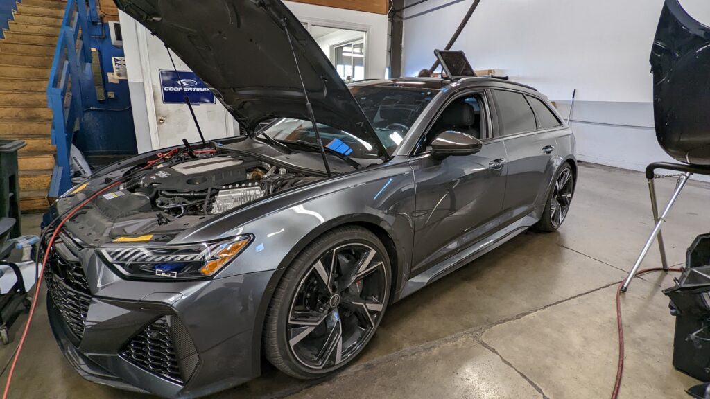 Auto repair shop in Southern Oregon with a gray European wagon on a lift, hood open, technician performing diagnostic testing with equipment connected in a clean, professional garage setting